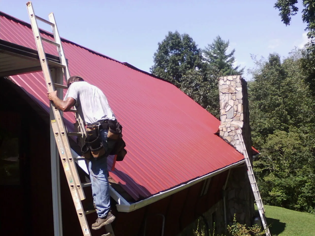 Roofer on a ladder beside a red metal roof and stone chimney — Soffit & Fascia Repair by Roofing Brothers in Trabuco Canyon